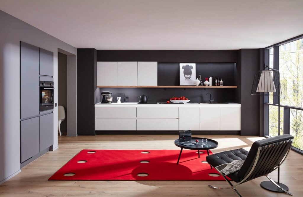 Moderne weiße Küchenzeile vor dunkler Rückwand, daneben eine graue Hochschrankwand mit Backofen, im Vordergrund ein roter Teppich und ein Lounge-Sessel