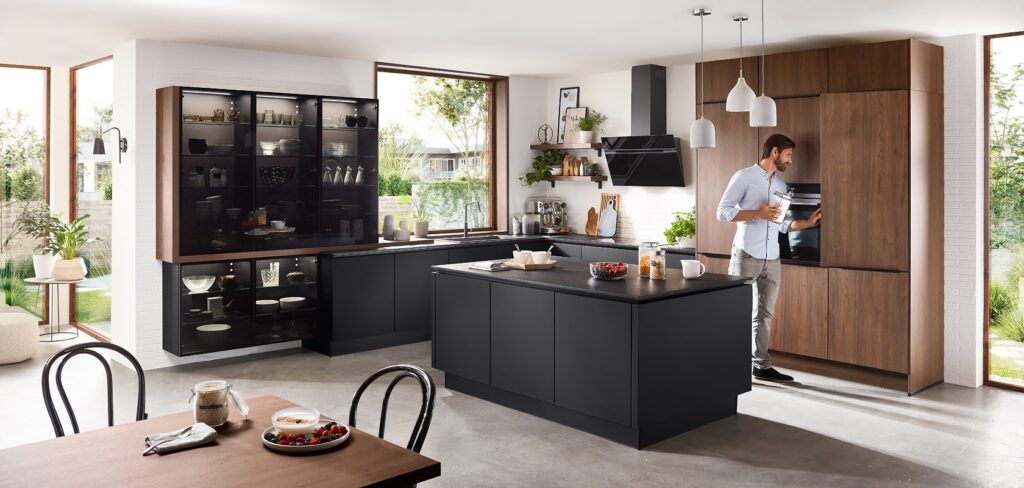 Offene Küche mit schwarzer Kochinsel, Hochschränken in Nussbaum-Optik und beleuchteter Glasvitrine; ein Mann nimmt etwas aus dem Backofen